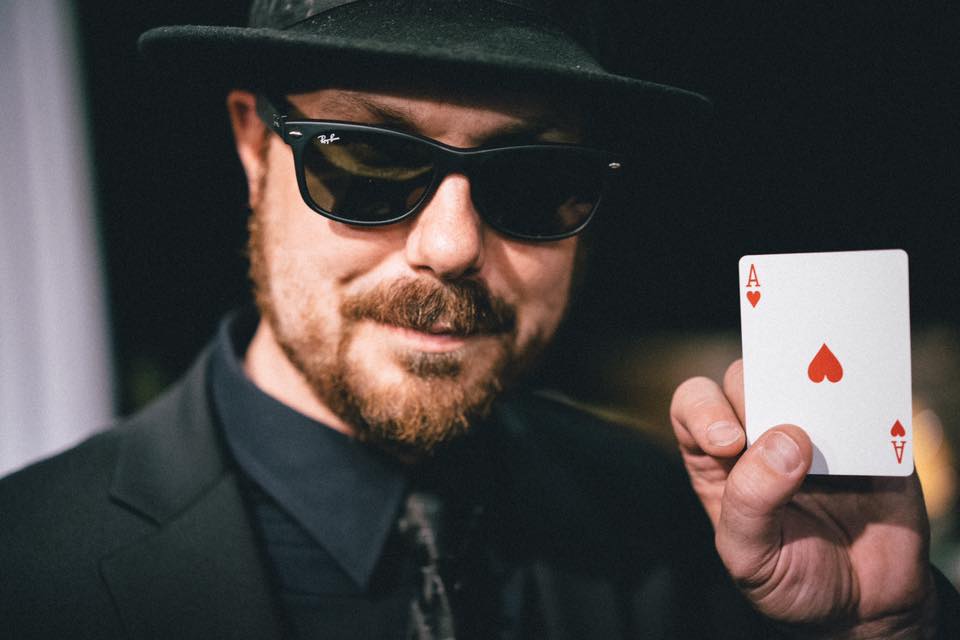Chad Allen wears a hat and holds up an Ace of hearts card.