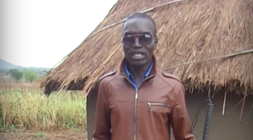 Francis Okello Oloya stands in front of a grass-roofed house.