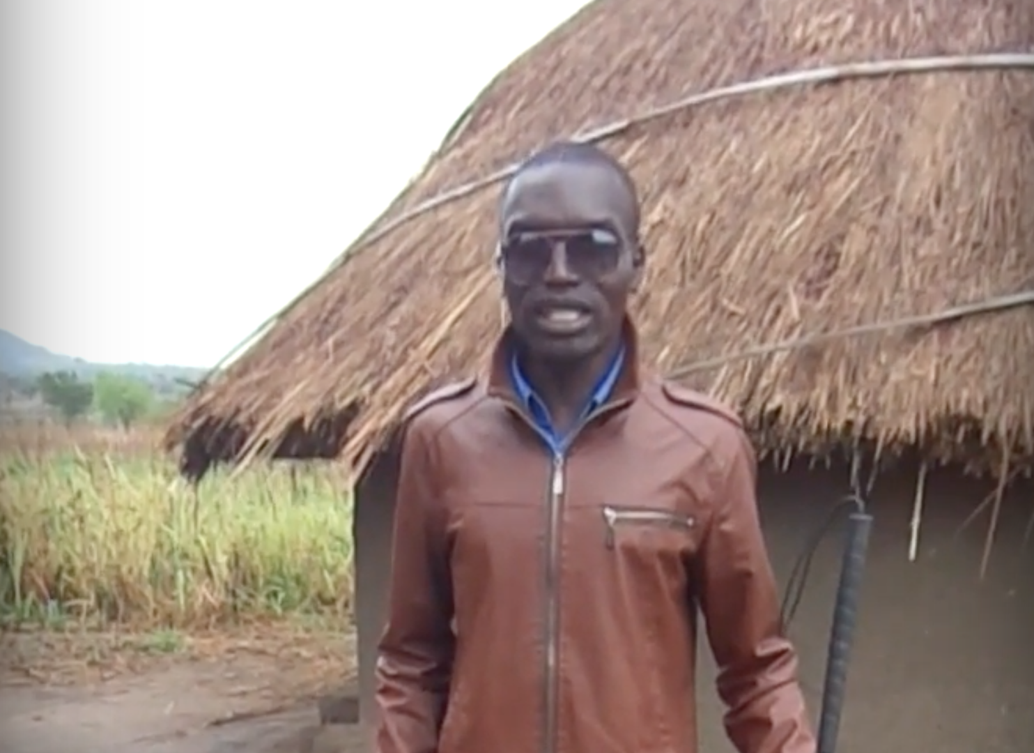 Francis Okello Oloya stands in front of a grass-roofed house.