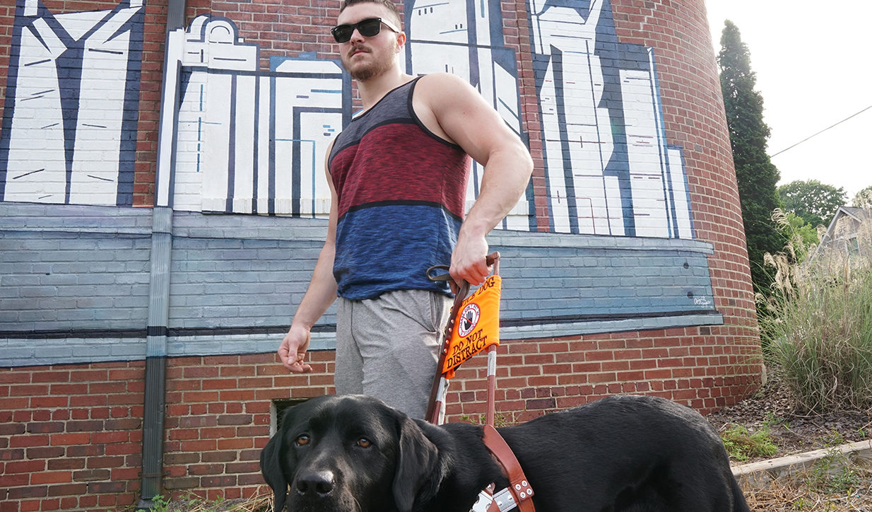 Justin Holland walks in front of a graffitied wall with his guide dog.