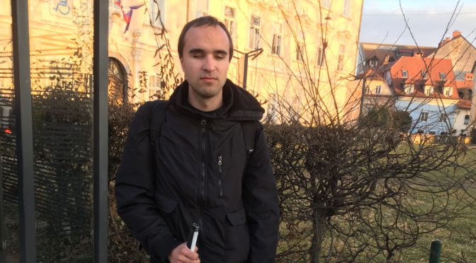 Miha Srebrnjak stands outside holding his white cane.